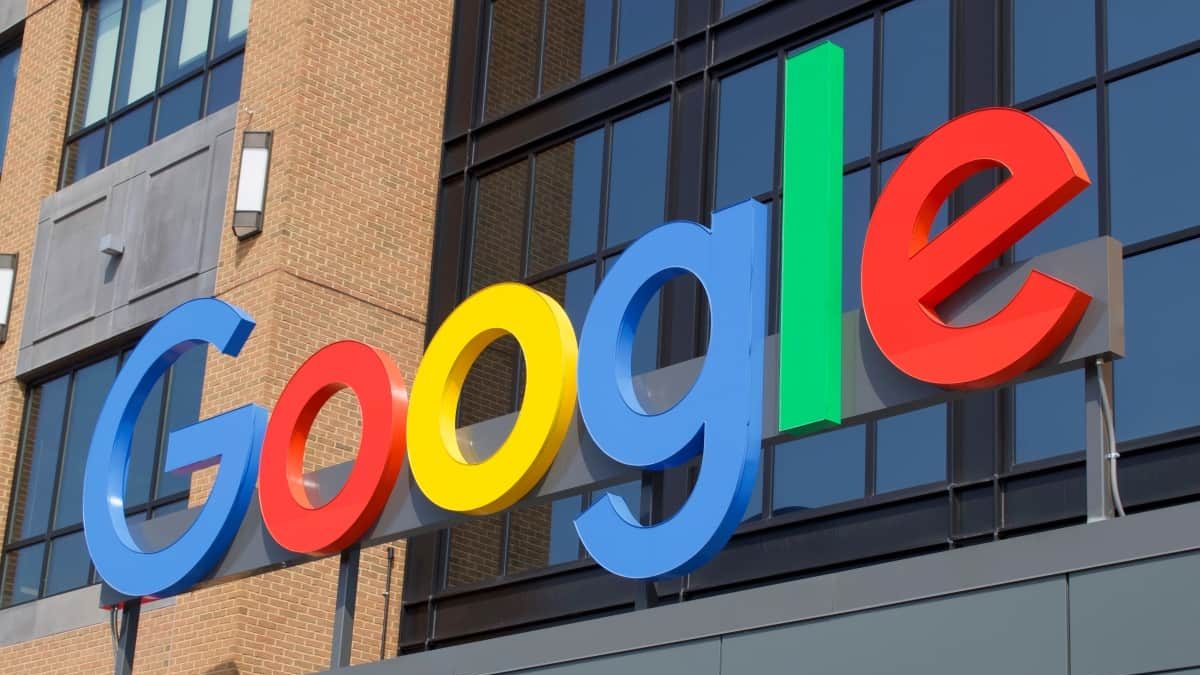 Logo colorido do Google em 3D fixado na fachada de um prédio com paredes de tijolos e janelas de vidro.