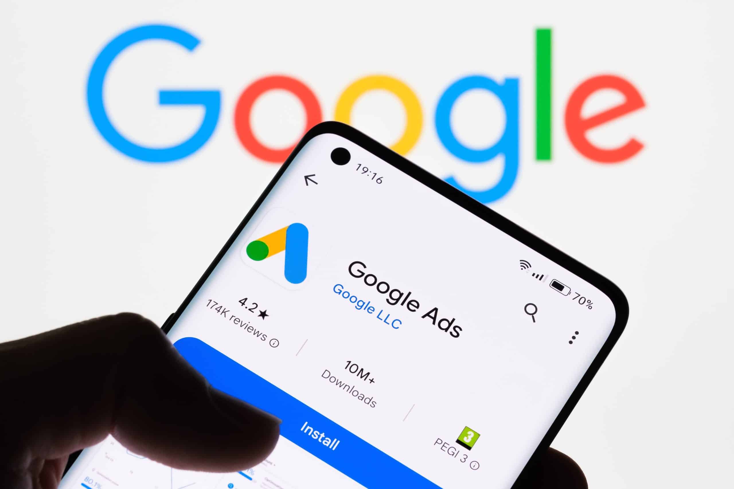 Mão segura smartphone exibindo o app Google Ads com 4.2 estrelas e 10M+ downloads. Logo Google colorido ao fundo.