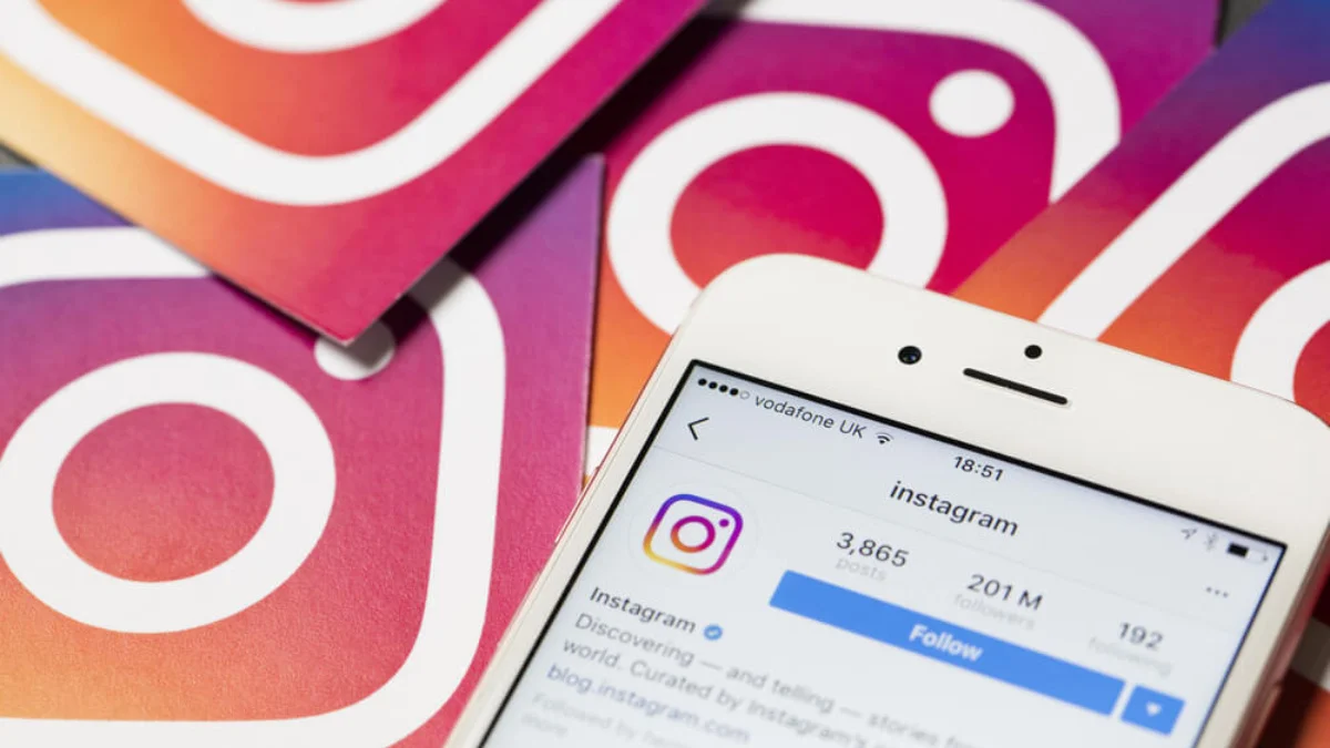 Smartphone branco com app Instagram aberto exibindo perfil com 201M seguidores, sobre logos coloridos do Instagram.
