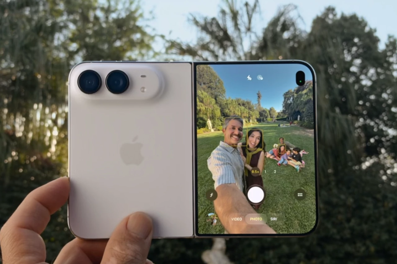 Mão segura smartphone dobrável branco com logo Apple e duas câmeras. Tela exibe selfie de casal e família em parque gramado.
