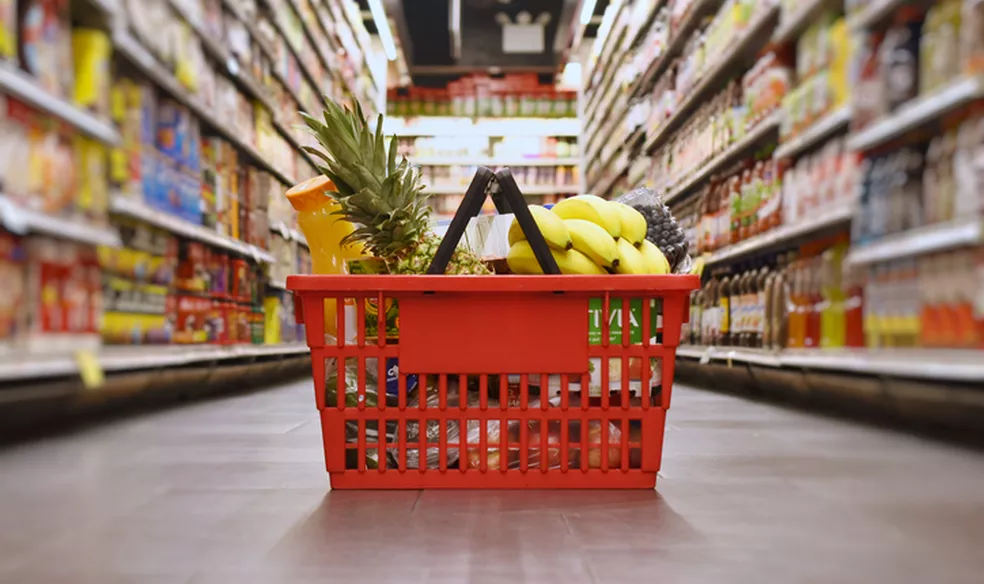 Cesta de compras vermelha cheia de frutas e garrafa amarela no corredor desfocado de um supermercado.