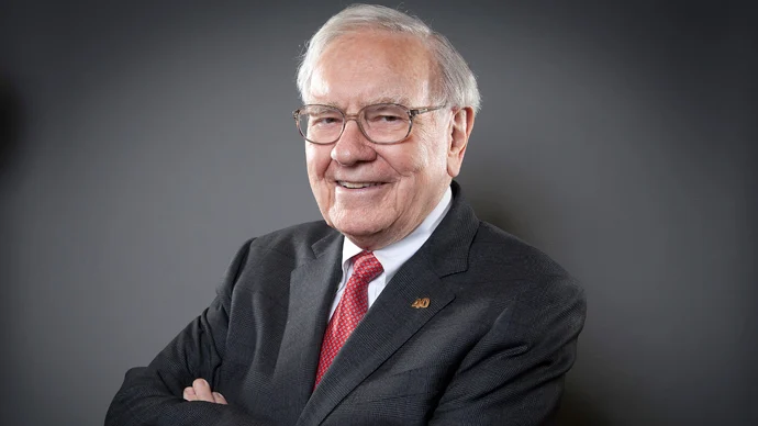 Warren Buffett, idoso de cabelos brancos, óculos, terno e gravata vermelha, sorrindo com braços cruzados. Fundo cinza.