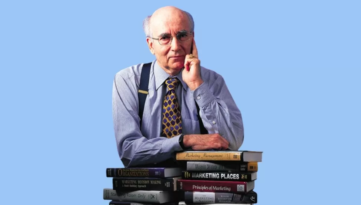 Philip Kotler, careca, de óculos e gravata, com a mão no rosto, apoiado em pilha de livros de marketing.